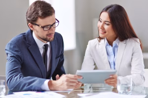 Two business professionals, a man in a suit and glasses and a woman in a white blazer, smiling as they review content on a tablet computer together at a glass desk.