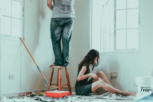 Couple painting and renovating a room together with paint tools and drop sheets around, illustrating a DIY home renovation scene